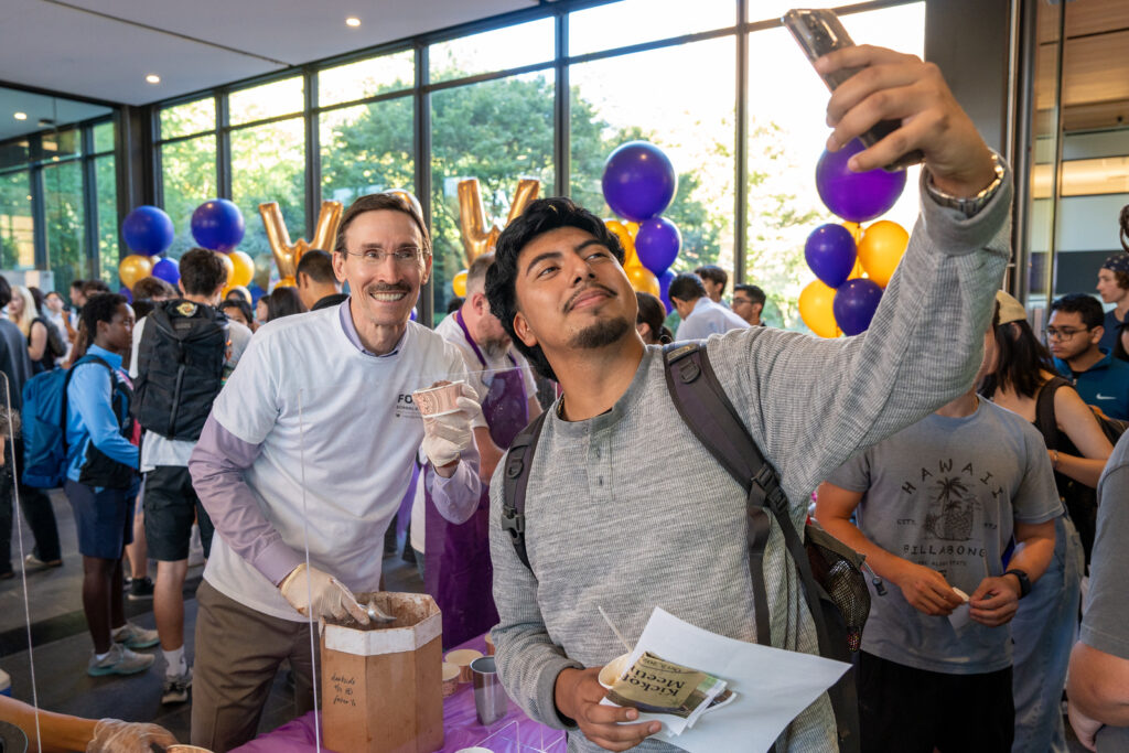 A man takes a selfie with a smiling individual at a lively event filled with people and colorful balloons.