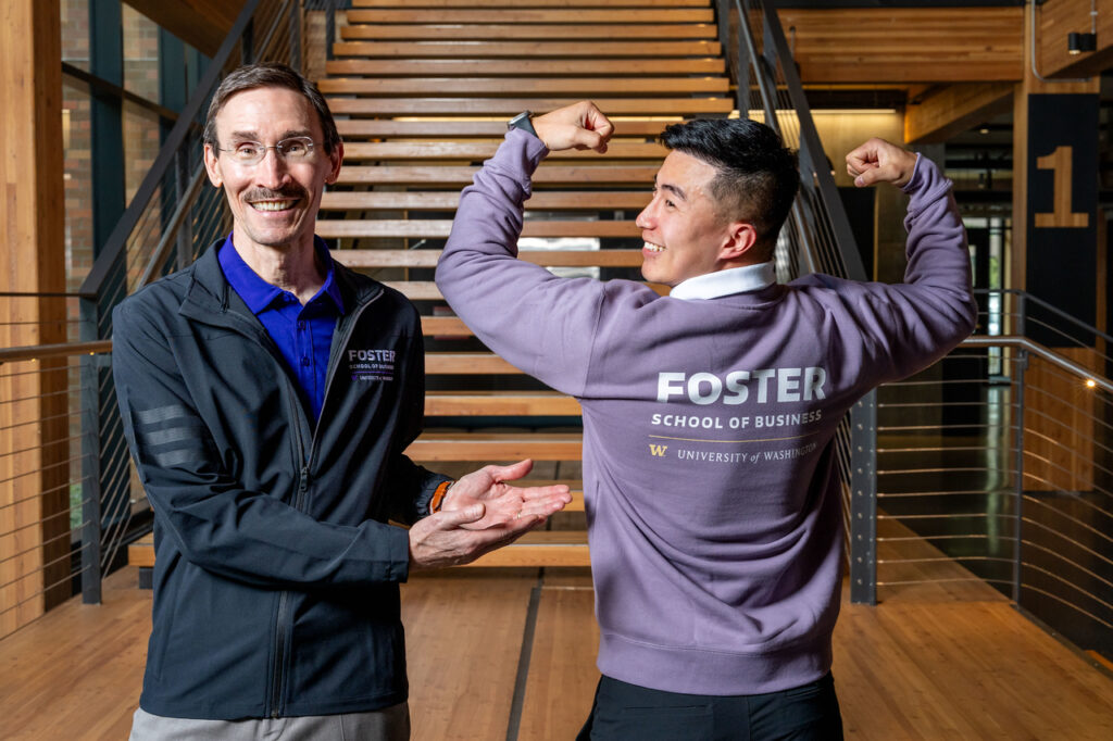 Two men pose in a modern building. One smiles and gestures, while the other flexes his muscles, both wearing Foster School of Business apparel.